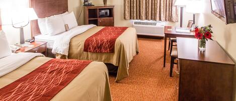 A hotel room with two beds, a desk, a chair, a TV, a lamp, a vase with flowers, and an air conditioning unit.
