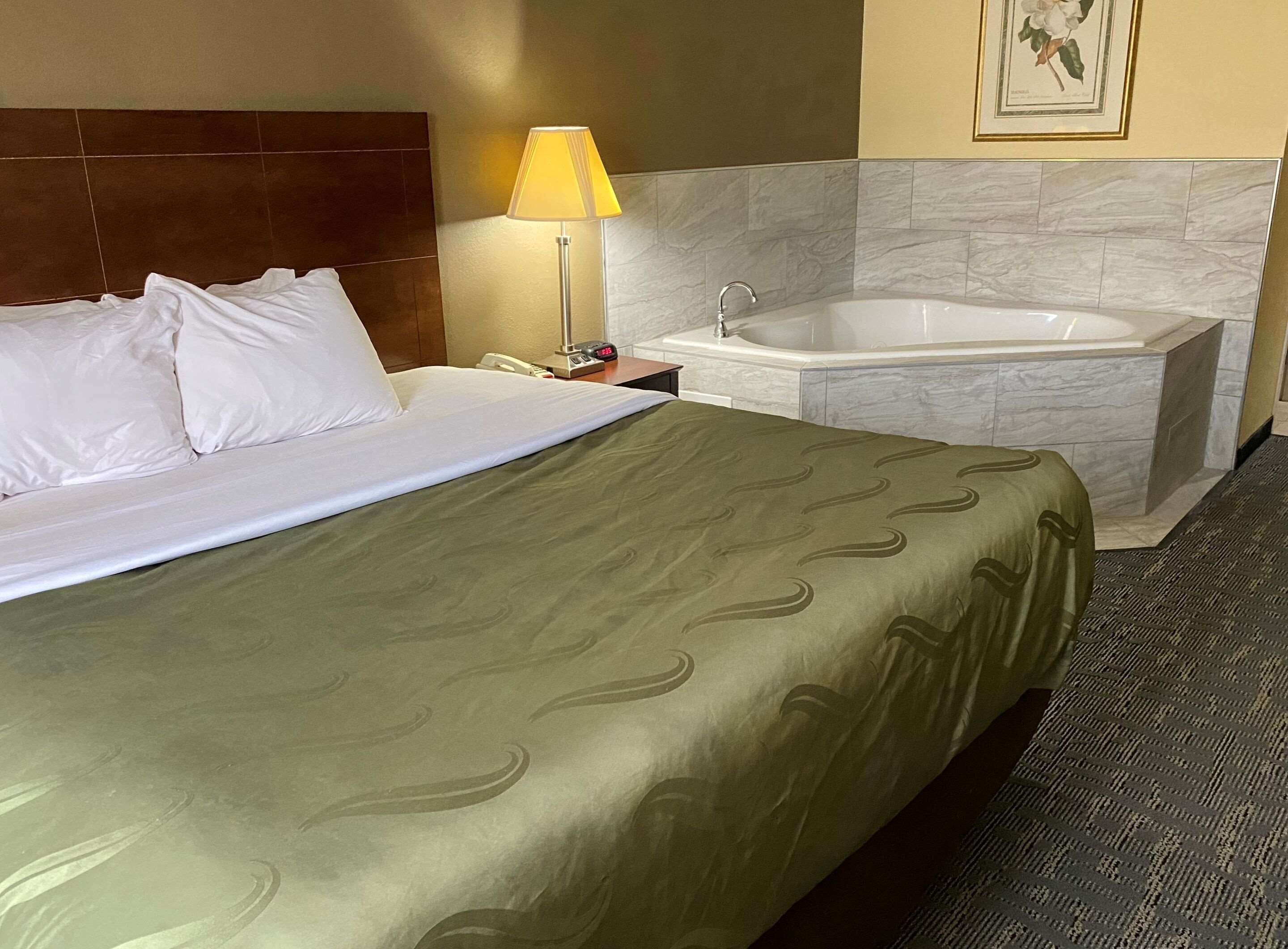 A hotel room with a bed, a nightstand, a lamp, and a bathtub.