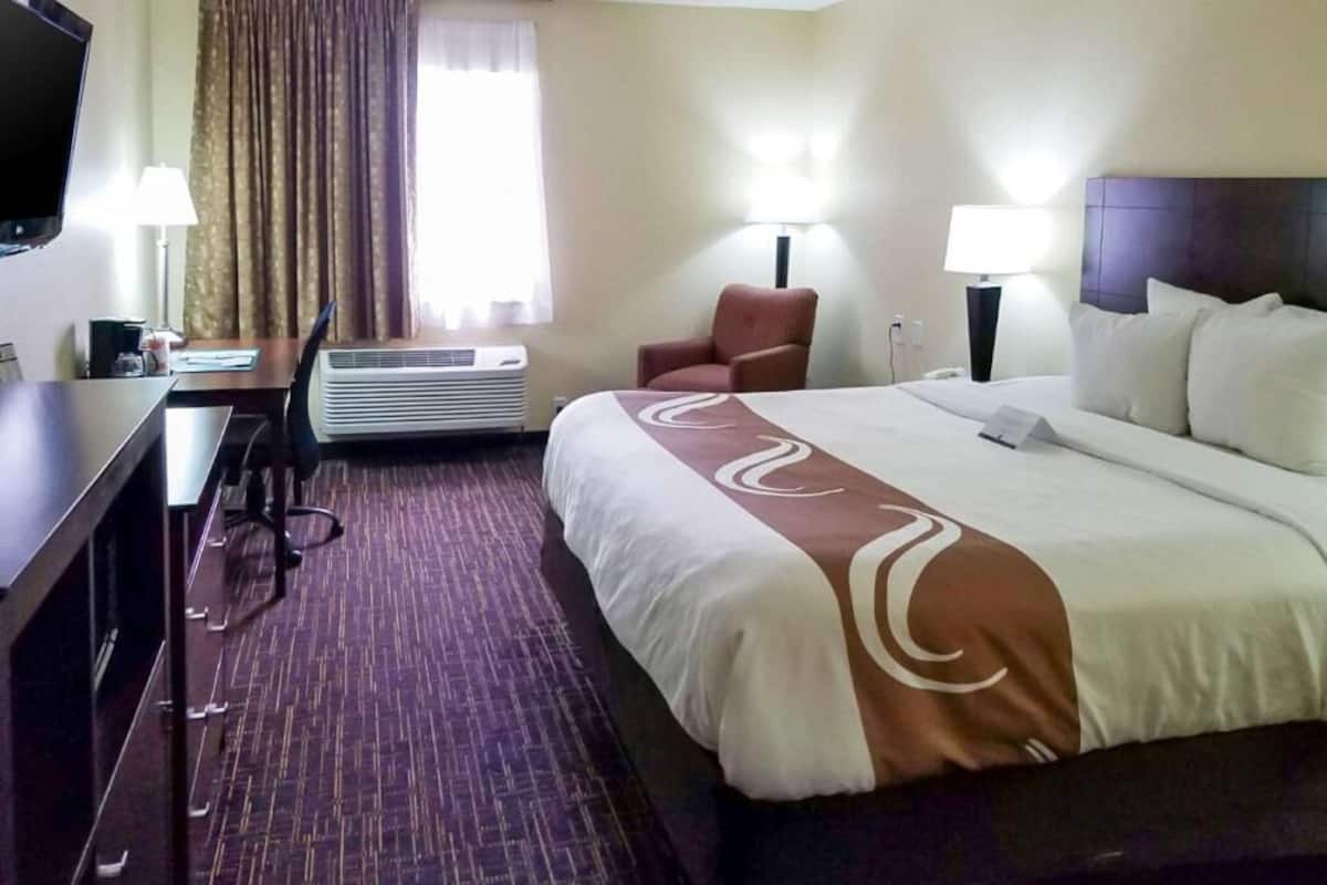 A hotel room with a bed, desk, chair, lamps, and an air conditioning unit.
