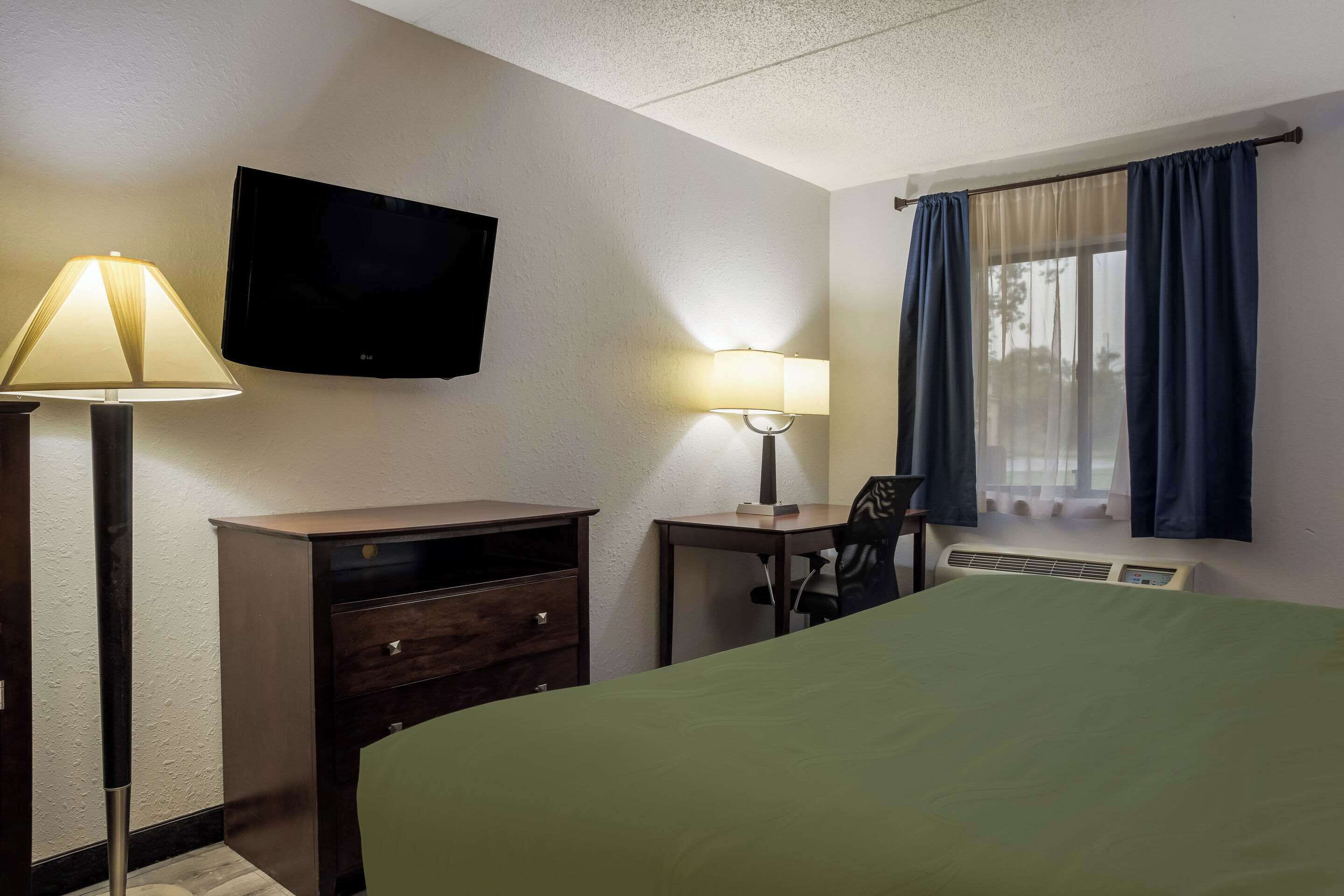 A hotel room with a bed, a desk with a chair, a TV mounted on the wall, a lamp, and a window with curtains.