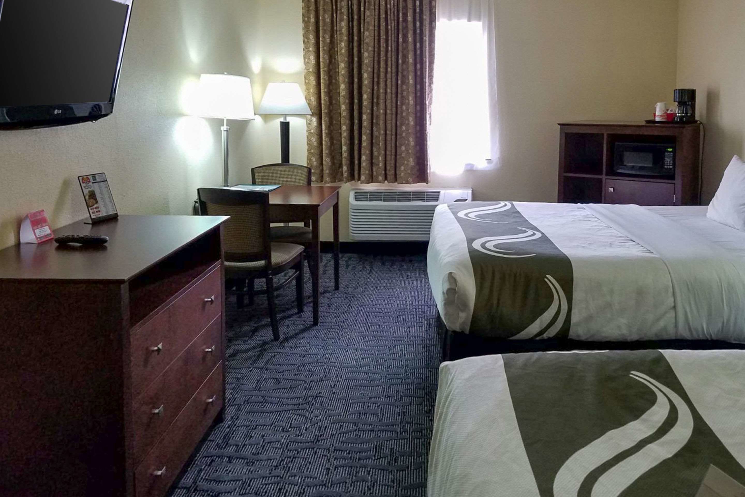 A hotel room with a bed, a desk with a chair, a television, a lamp, a microwave, and a dresser.