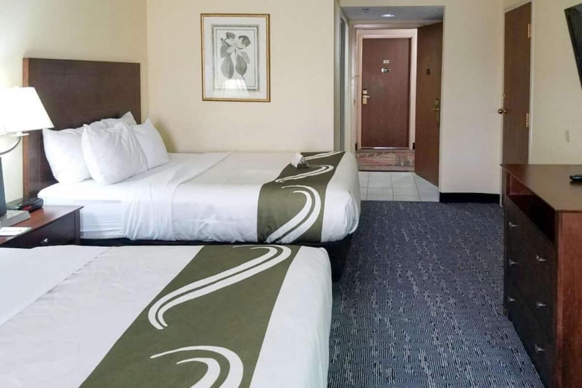 A hotel room with two beds, a desk, a lamp, a framed picture on the wall, and a door leading to another room.