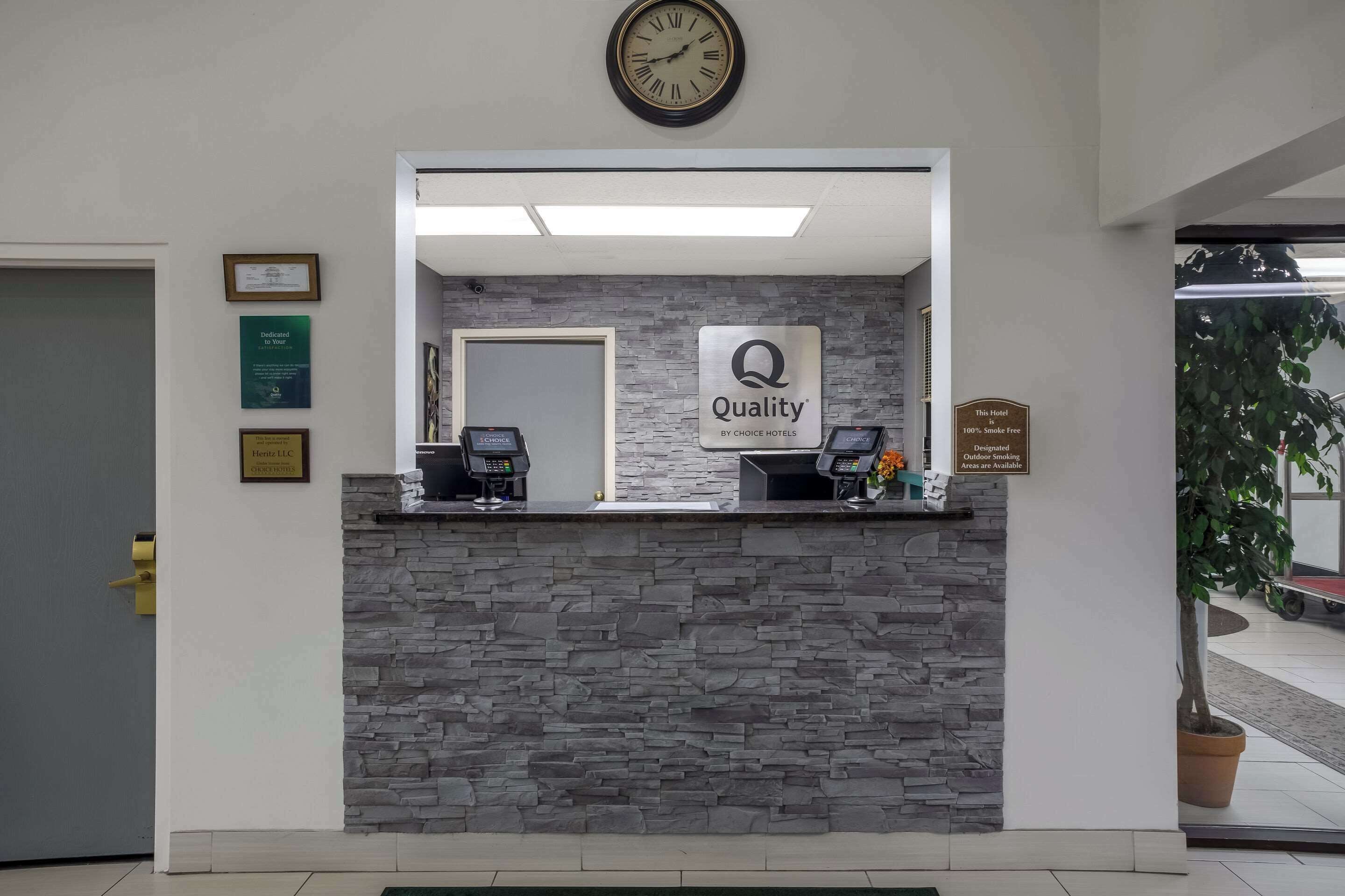A hotel reception desk with a clock, a sign reading "Quality by Choice Hotels," and a plaque indicating "100% Smoke Free."