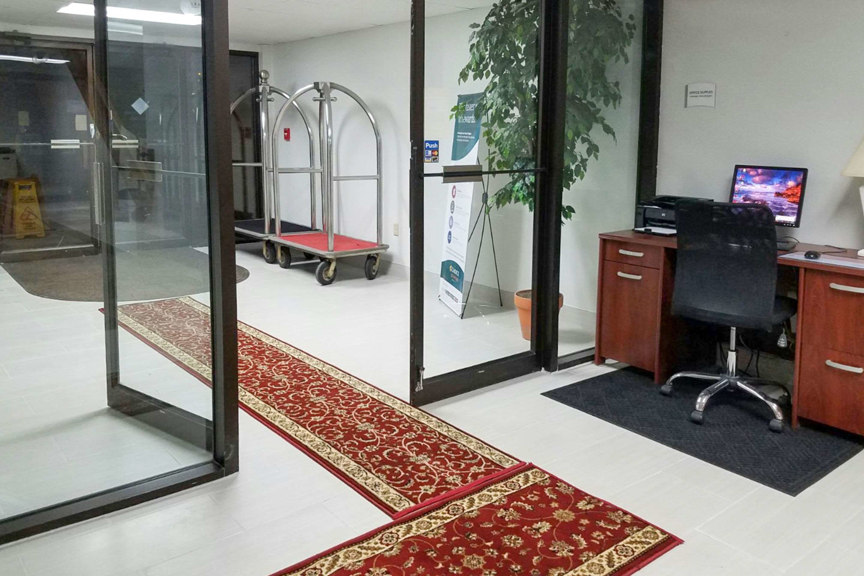 A modern office space with a desk, chair, computer, and a red carpet leading to a glass door.