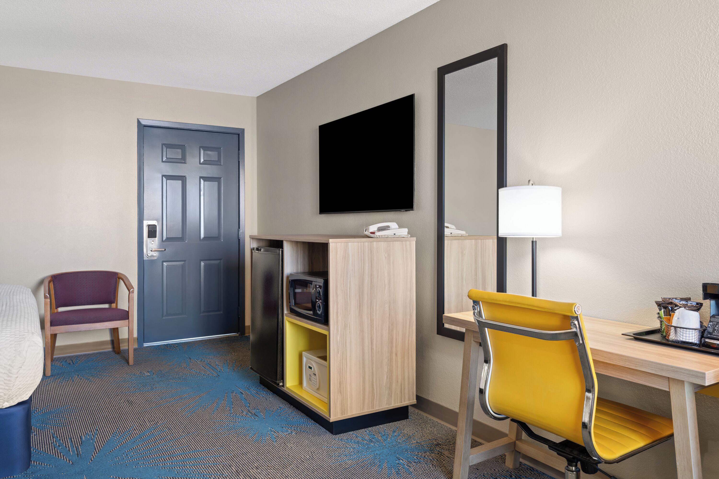Een hotelkamer met een bed, een bureau met stoel, een televisie, een deur en een klein keukenhoekje.