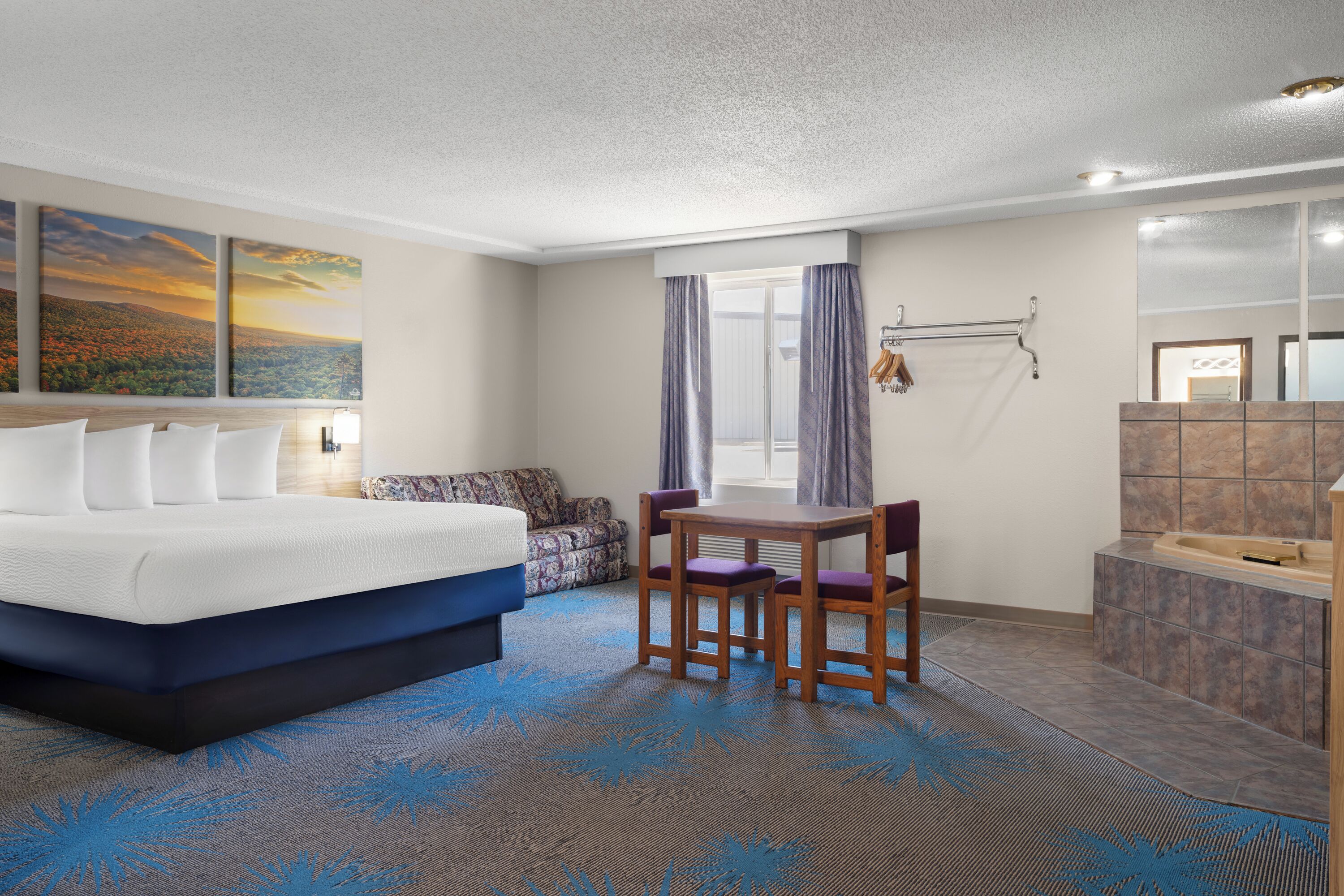 A hotel room with a bed, a table with chairs, a small sofa, a window with curtains, a bathroom with a bathtub, and a large mirror.