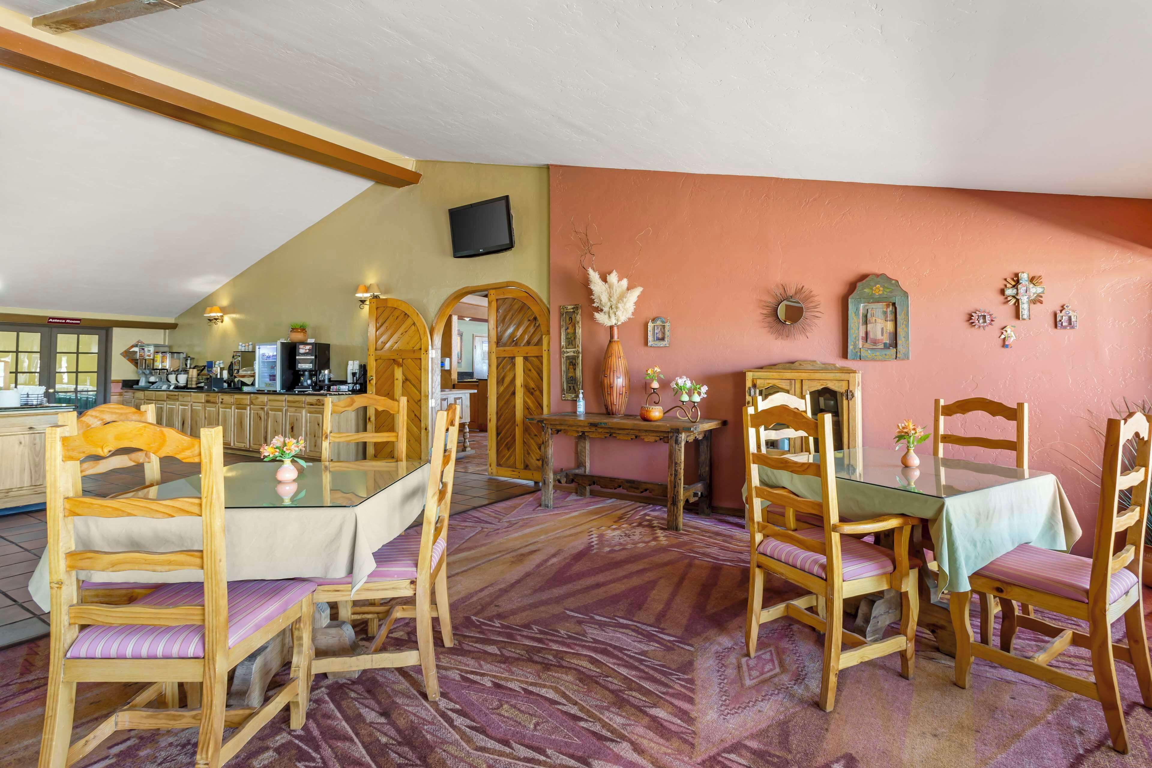Un coin repas avec des chaises en bois et une table en verre, une cuisine à l’arrière-plan et un téléviseur fixé au mur.