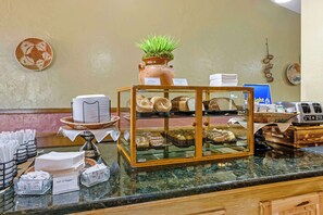 Uma área de buffet com diversos produtos de pastelaria, um espaço para café e uma planta decorativa.