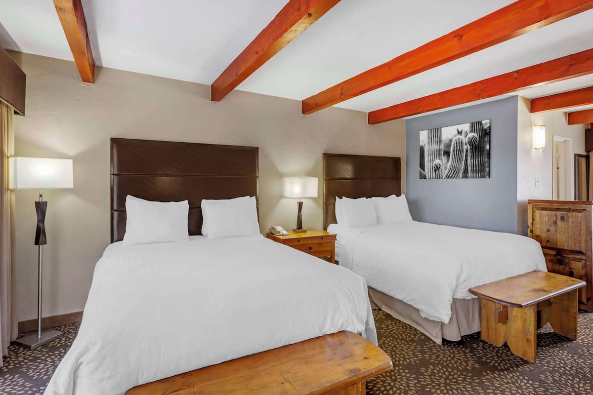 A hotel room with two beds, wooden headboards, a nightstand with a lamp, a small wooden table, and a wall-mounted artwork.