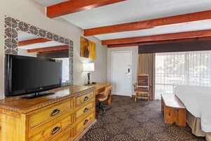 A hotel room with a wooden dresser, a flat-screen TV, a desk with a chair, a bed, and a patterned carpet.
