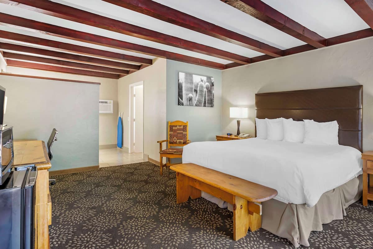 A hotel room with a large bed, a wooden bench, a desk with a computer, a chair, and a wall-mounted artwork.