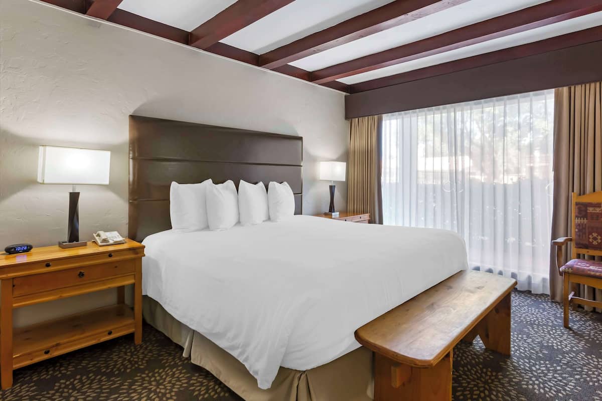 A hotel room with a large bed, wooden nightstands, a bench, and a view of the outdoors through sheer curtains.