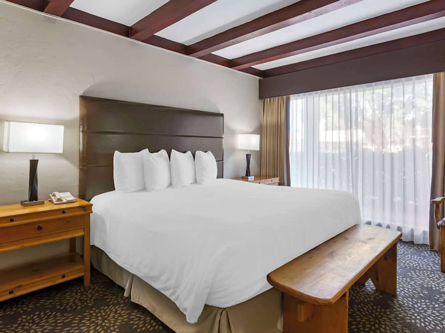 A hotel room with a large bed, wooden nightstands, a bench, and a view of the outdoors through sheer curtains.