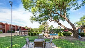 A patio with a table and chairs, a large tree, a fountain, and a pool in the background.
