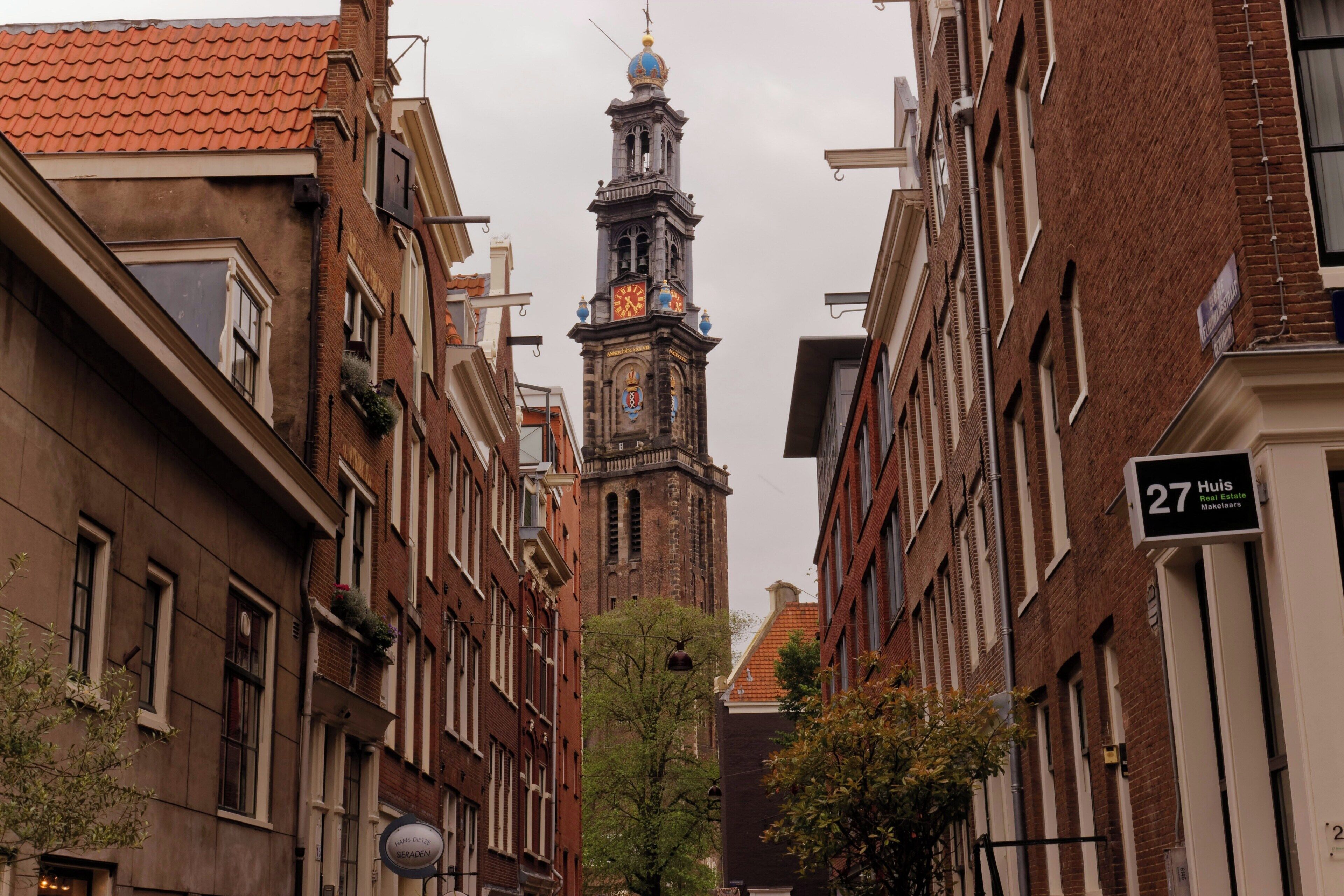 Una calle estrecha con arquitectura tradicional europea, que destaca una imponente torre del reloj y un edificio con un letrero que indica una oficina de bienes raíces.