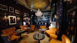 A bar area with orange upholstered seating, round tables, and a chandelier.