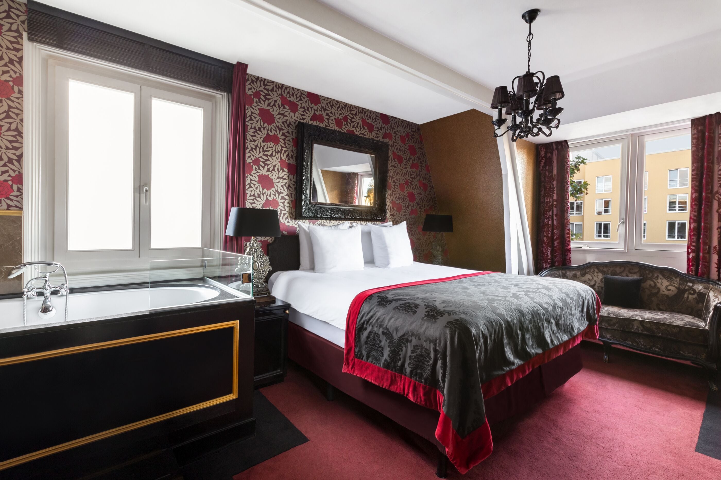 A hotel room with a large bed, a vanity with a mirror, a chandelier, and a view of buildings through the window.
