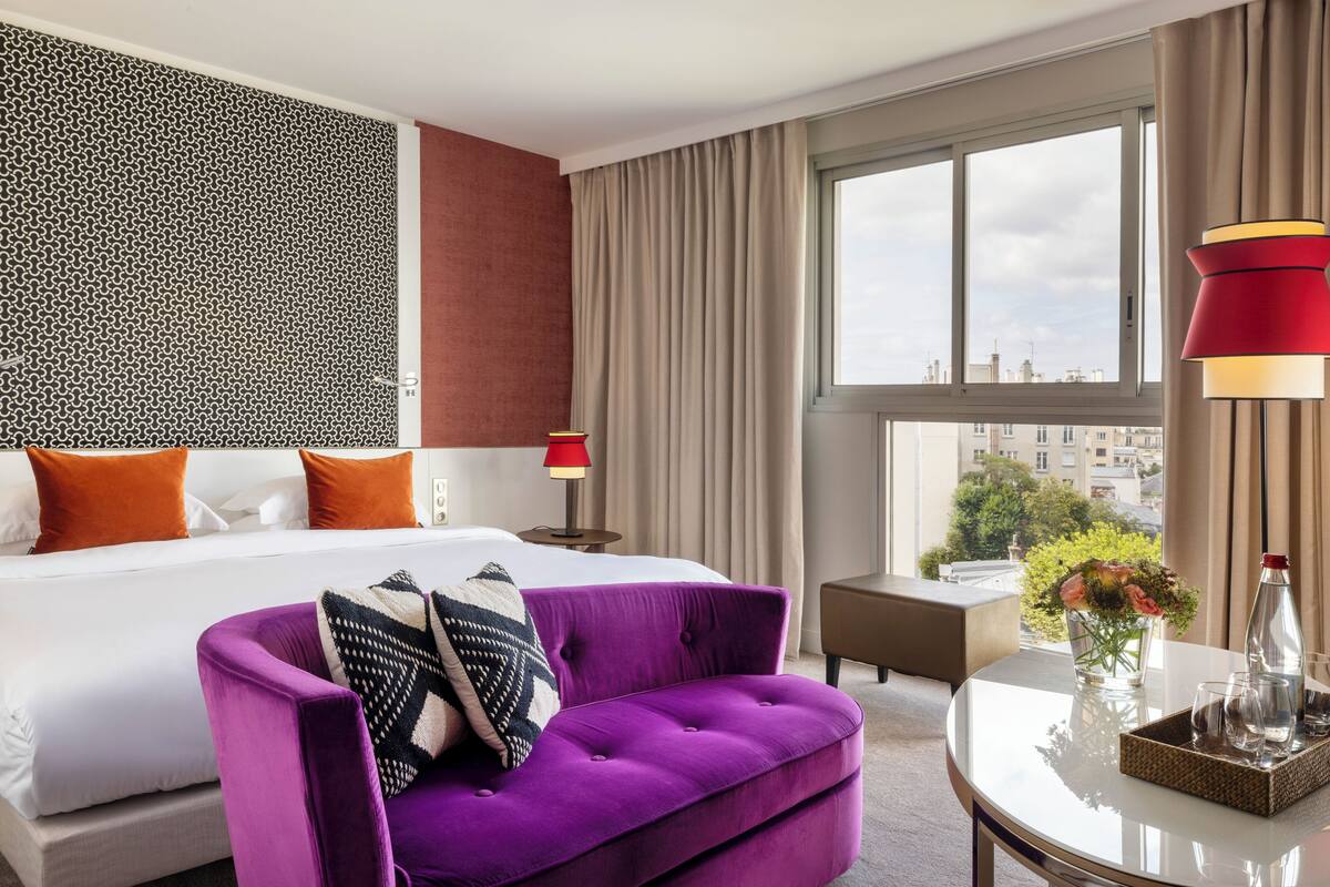 A hotel room with a large bed, a purple armchair, a small table with a vase of flowers, and a view of the city through the window.