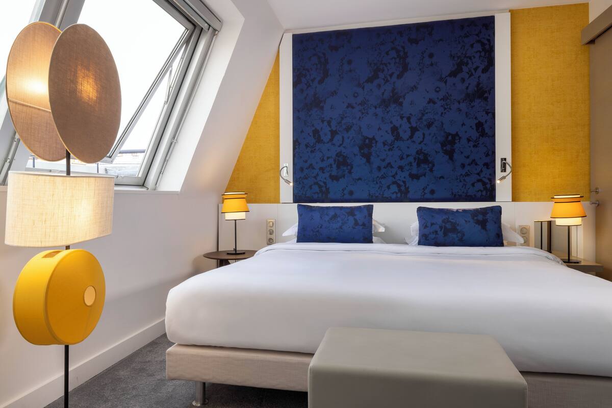 A modern hotel room with a large bed, two bedside tables with lamps, a large window, and a blue patterned wall.