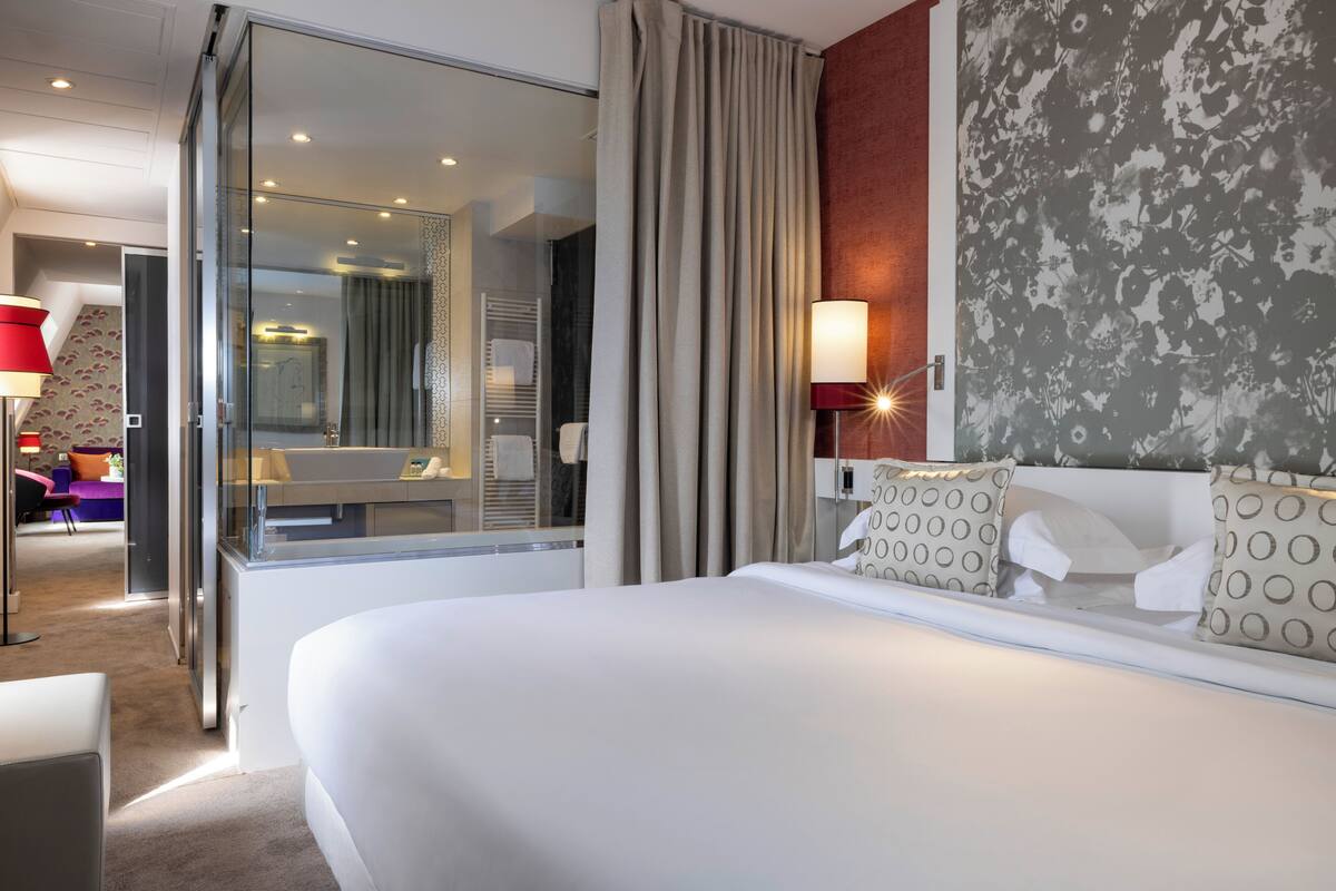A modern hotel room with a large bed, a glass door leading to a bathroom, a sofa, and a lamp.