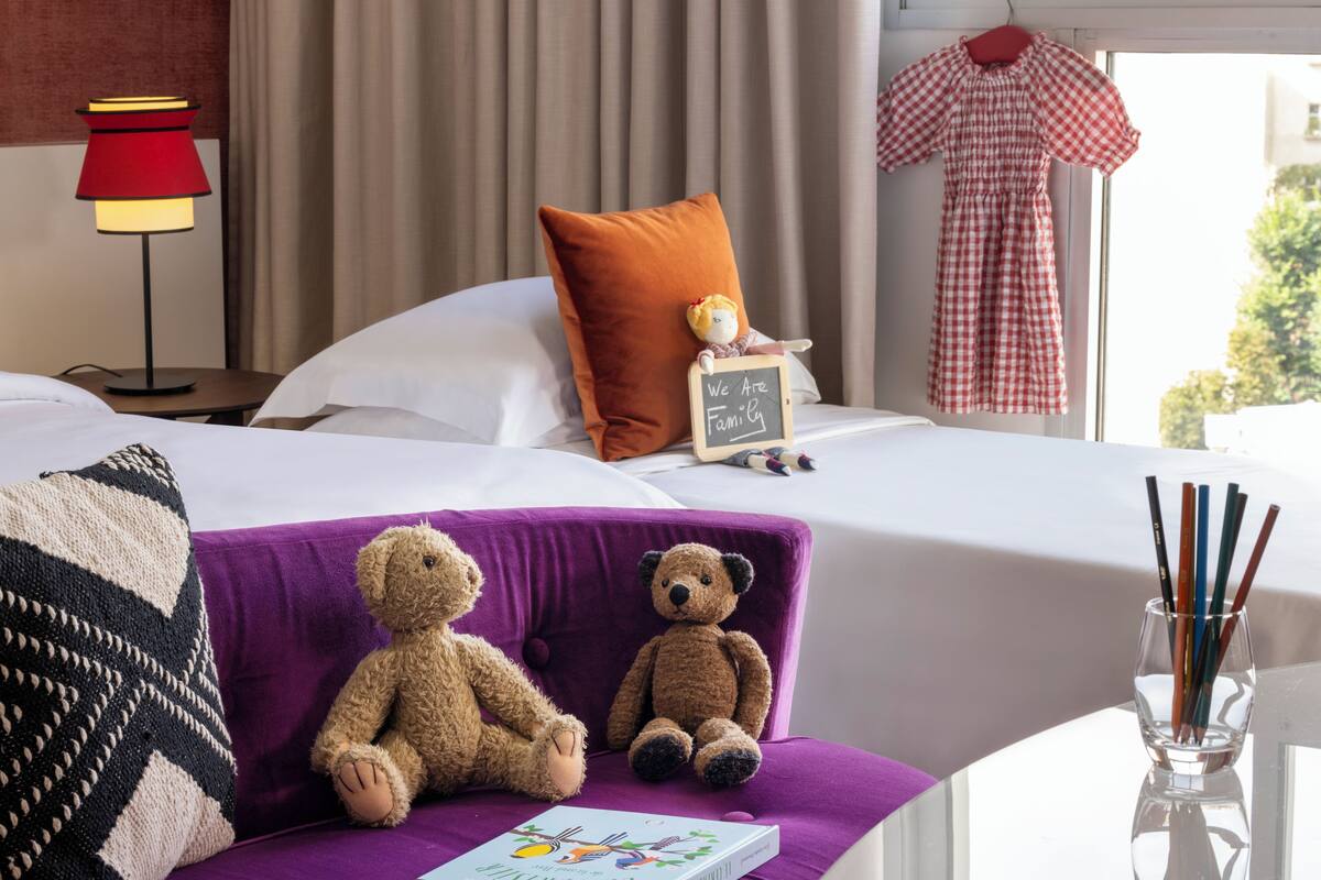 A hotel room with a bed, a purple sofa, two teddy bears, a small chalkboard sign, a red lamp, and a window with a view.