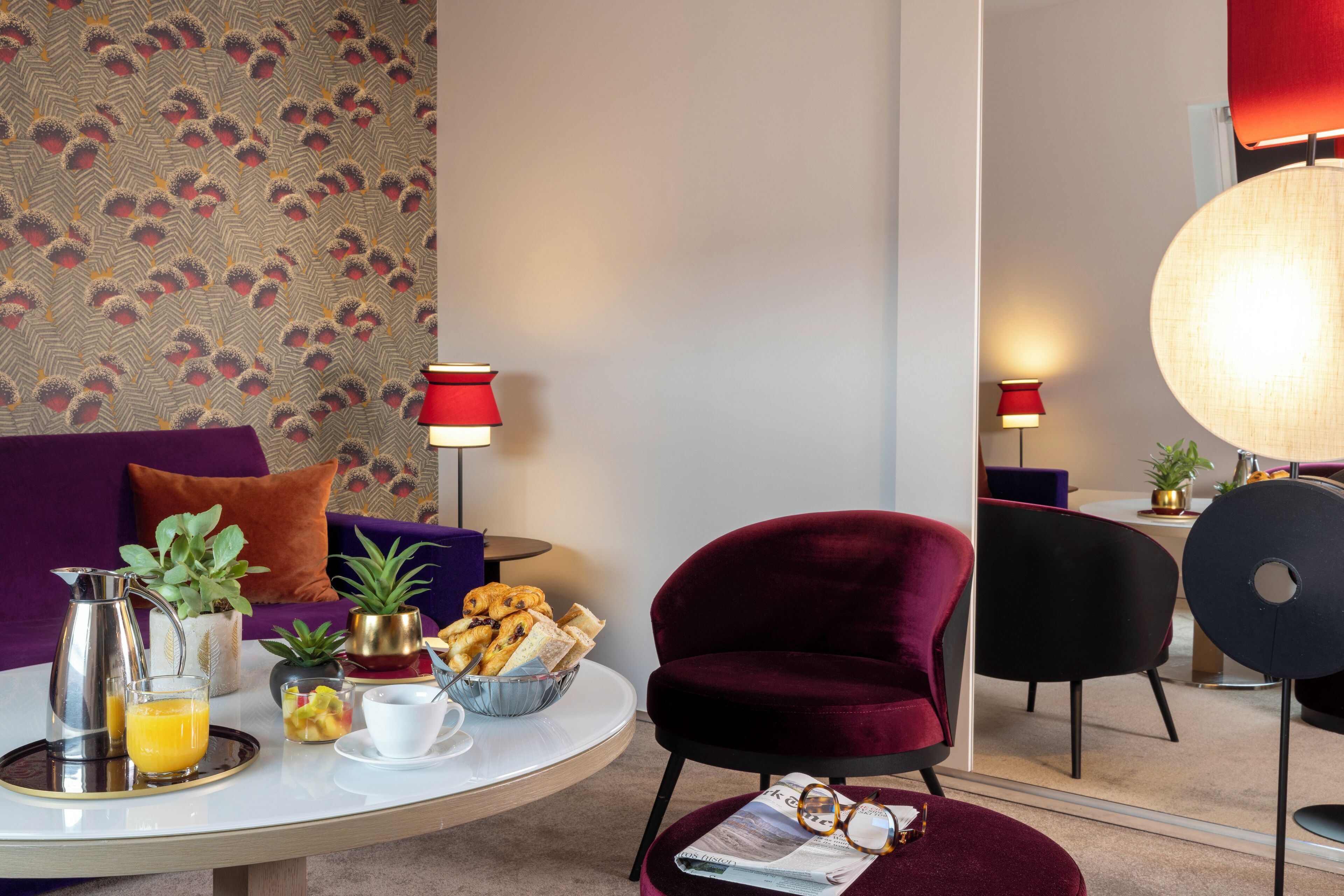 Una sala moderna con un sofá púrpura, un sillón rojo, una mesa de centro redonda con comida para el desayuno y un espejo grande que refleja la habitación.