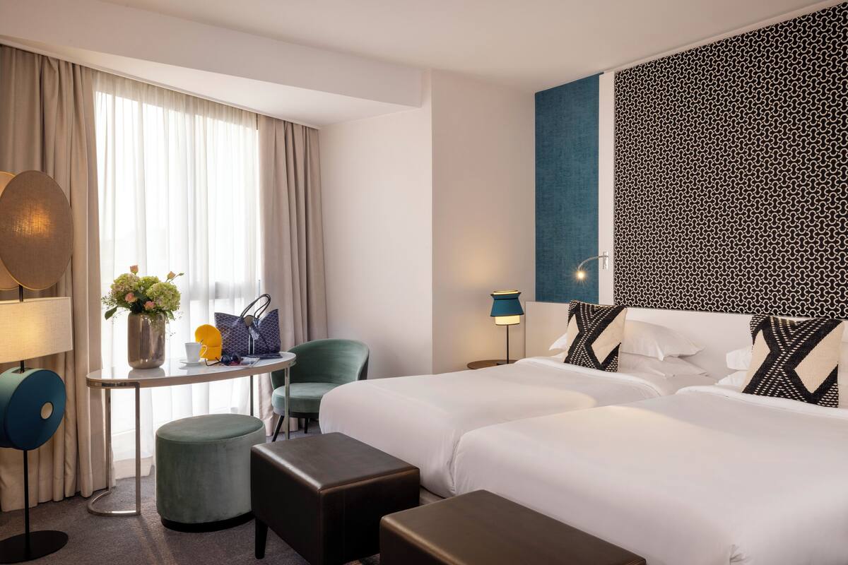 A hotel room with two beds, a desk with a lamp, a chair, and a small table with a vase of flowers.