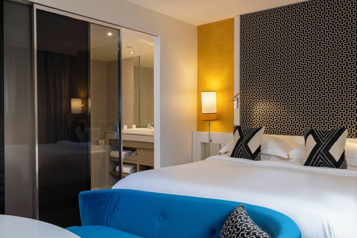 A modern hotel room with a large bed, a blue sofa, a glass door leading to a bathroom, and a patterned wall.