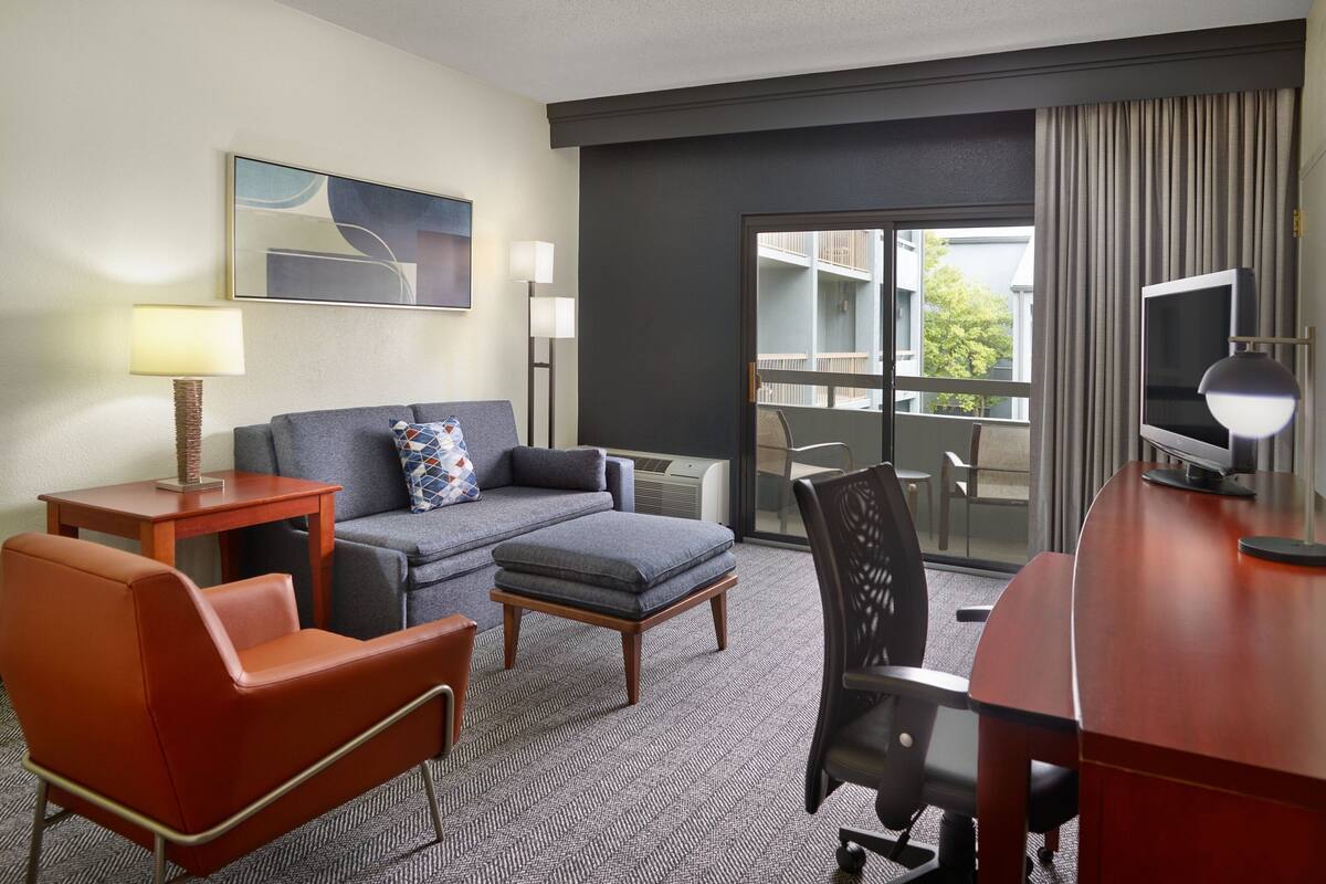 A hotel room with a sofa, armchair, ottoman, desk, TV, and a balcony with a view.