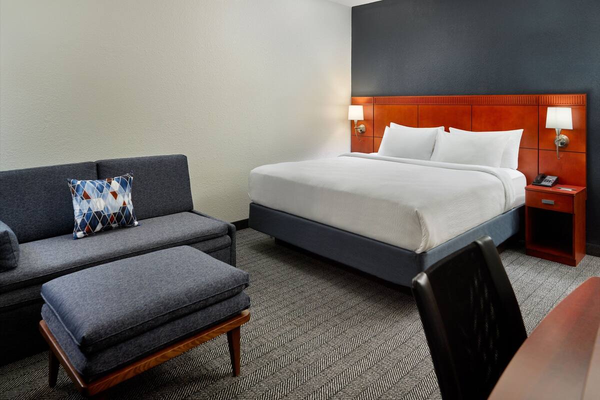 A hotel room with a bed, a sofa, an ottoman, a nightstand, and a desk.