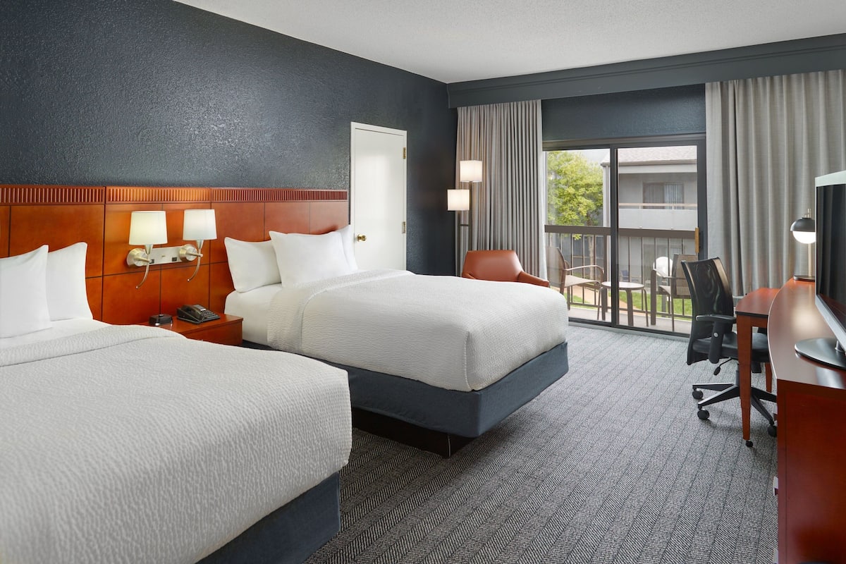 A hotel room with two beds, a desk, a chair, a TV, and a balcony with a view.