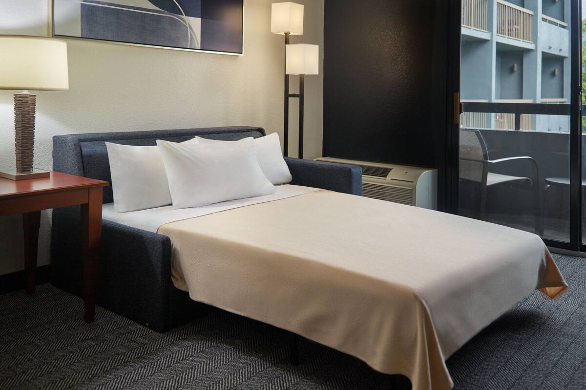 A hotel room with a bed, a nightstand, a lamp, and a view of an outdoor balcony.