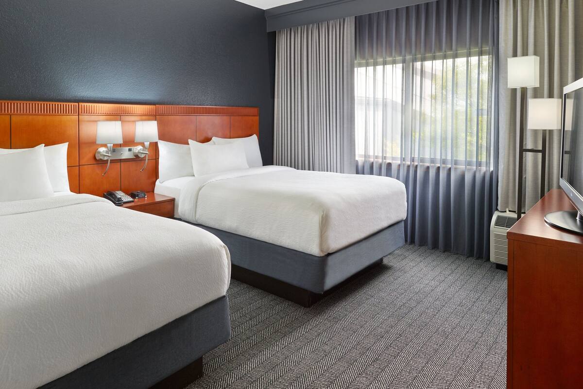 A hotel room with two beds, a wooden headboard, a flat-screen TV, and a large window with curtains.