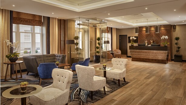 A hotel lobby with a reception desk, seating area with armchairs and sofas, and a large window with curtains.