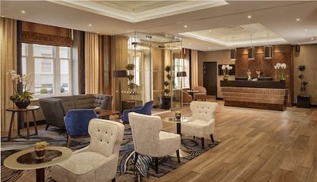 A hotel lobby with a reception desk, seating area with armchairs and sofas, and a large window with curtains.