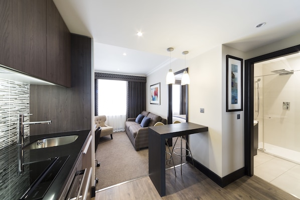 A modern kitchen with a built-in stove, a dining area with a table and chairs, a sofa, and a glass shower enclosure. - The Westbourne Hyde Park (London)