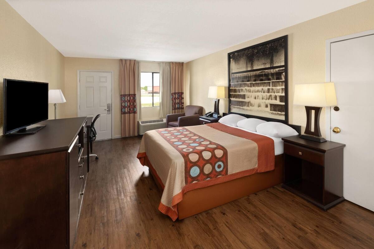 A hotel room with a bed, a desk with a TV, a chair, a nightstand, and a wall-mounted lamp.