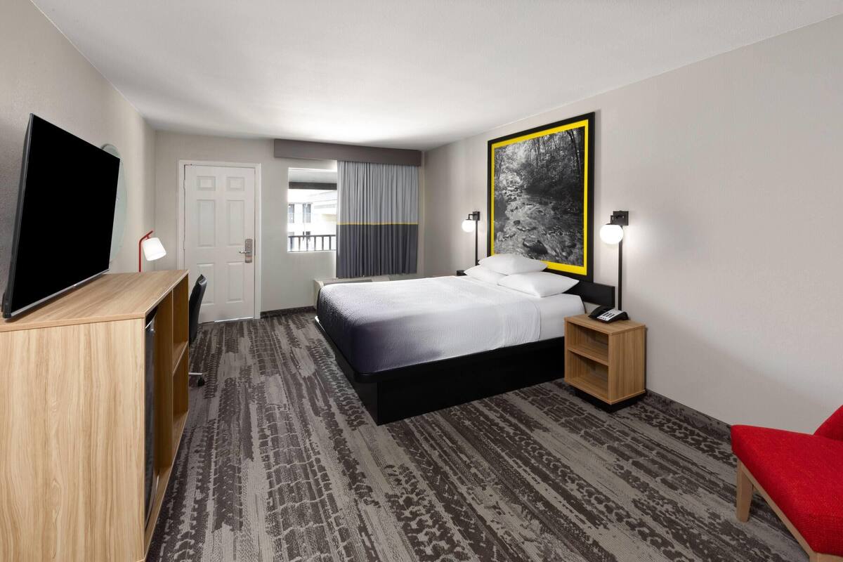 A hotel room with a bed, a TV, a desk, a chair, a bedside table with a lamp, and a large framed picture on the wall.