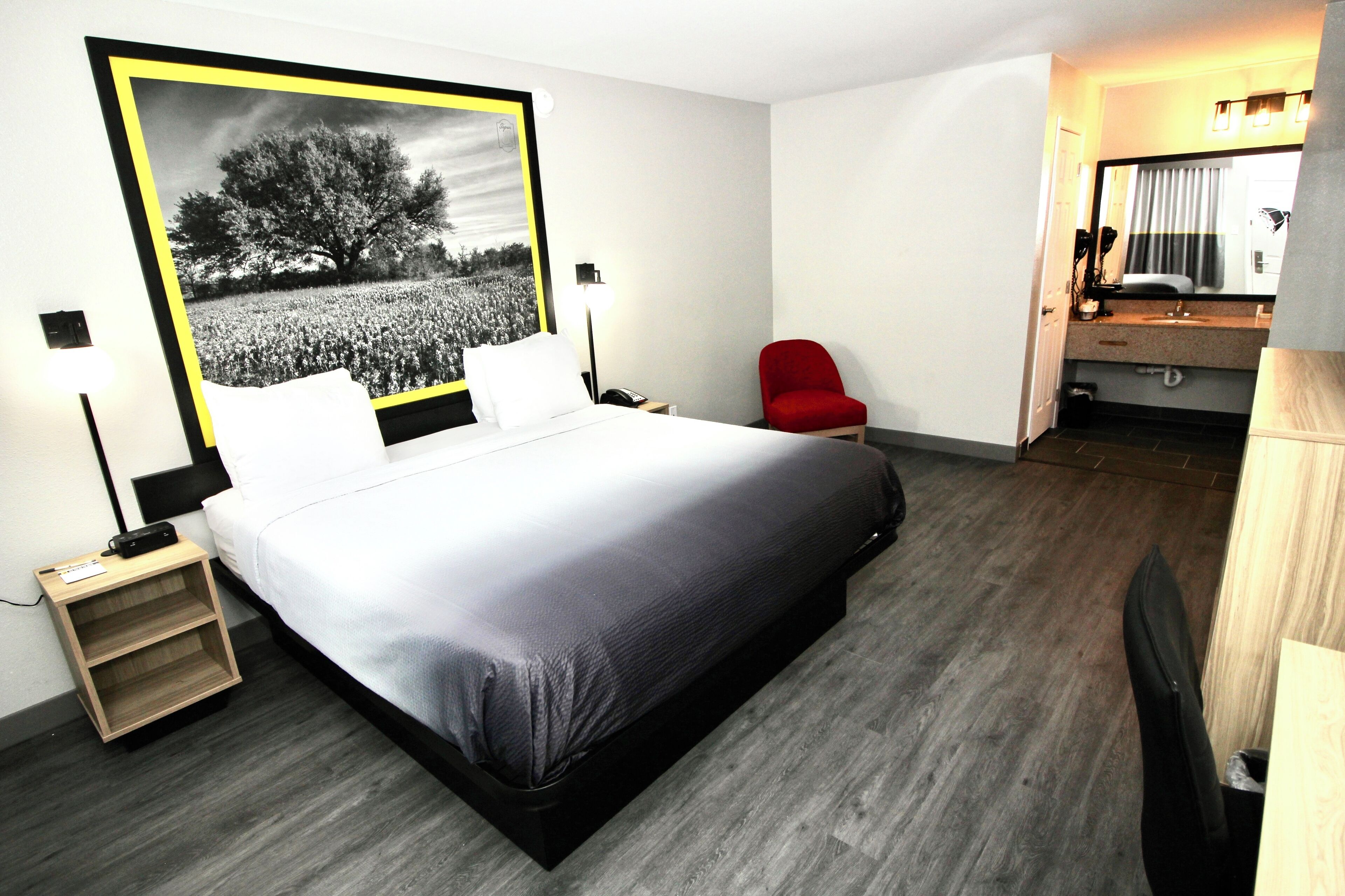 A modern hotel room with a large bed, a red chair, a desk with a lamp, and a bathroom visible through an open door.