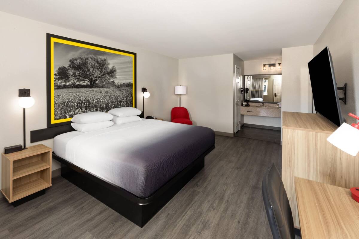 A modern hotel room with a large bed, bedside lamps, a red chair, a wooden nightstand, a TV, and a mirror.