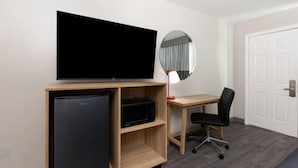 Habitación de hotel con televisión de pantalla plana, refrigerador, microondas, escritorio con silla, espejo redondo y puerta.