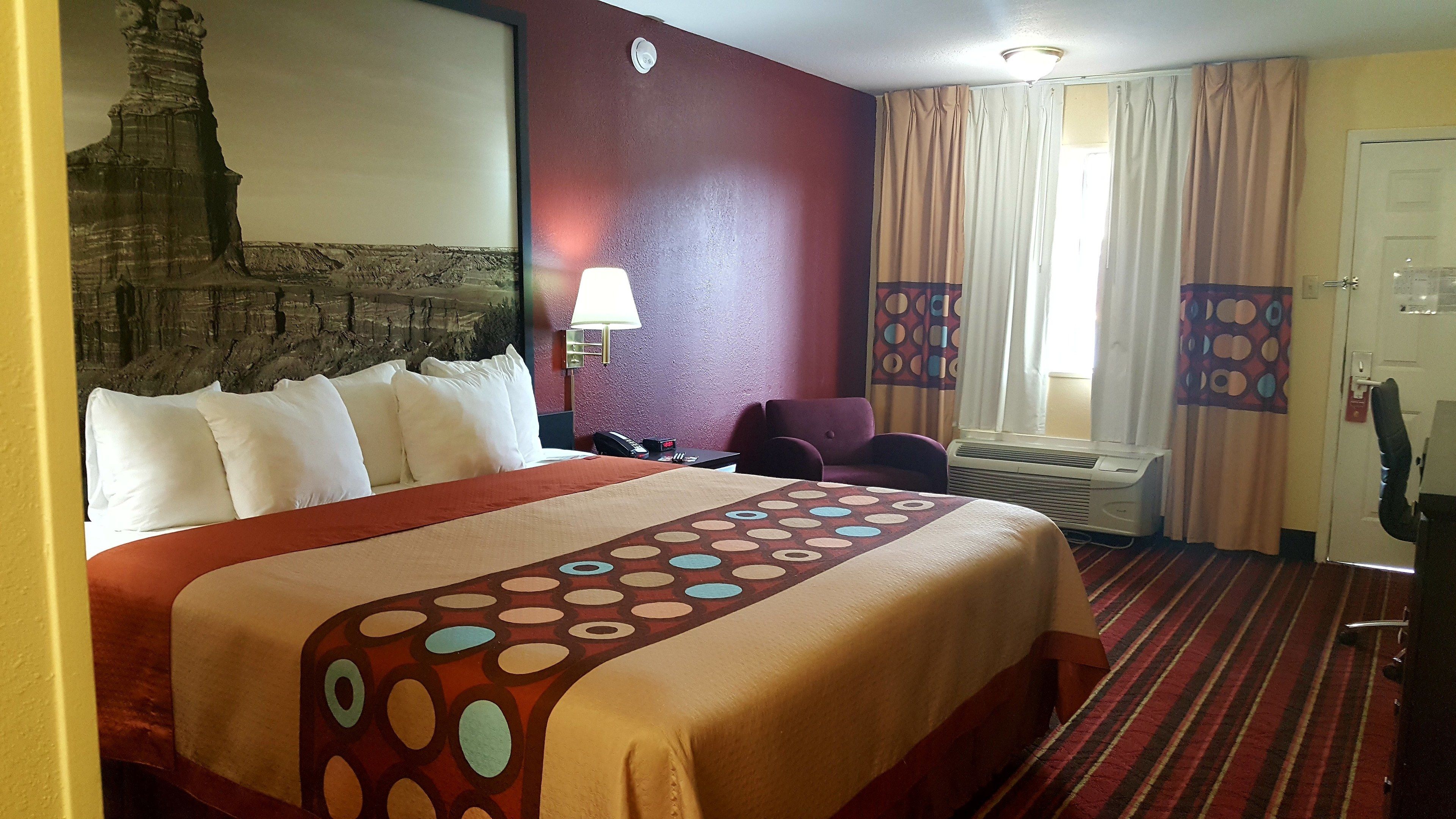Une chambre d’hôtel avec un grand lit, un bureau, une chaise, une lampe fixée au mur et une fenêtre avec des rideaux.