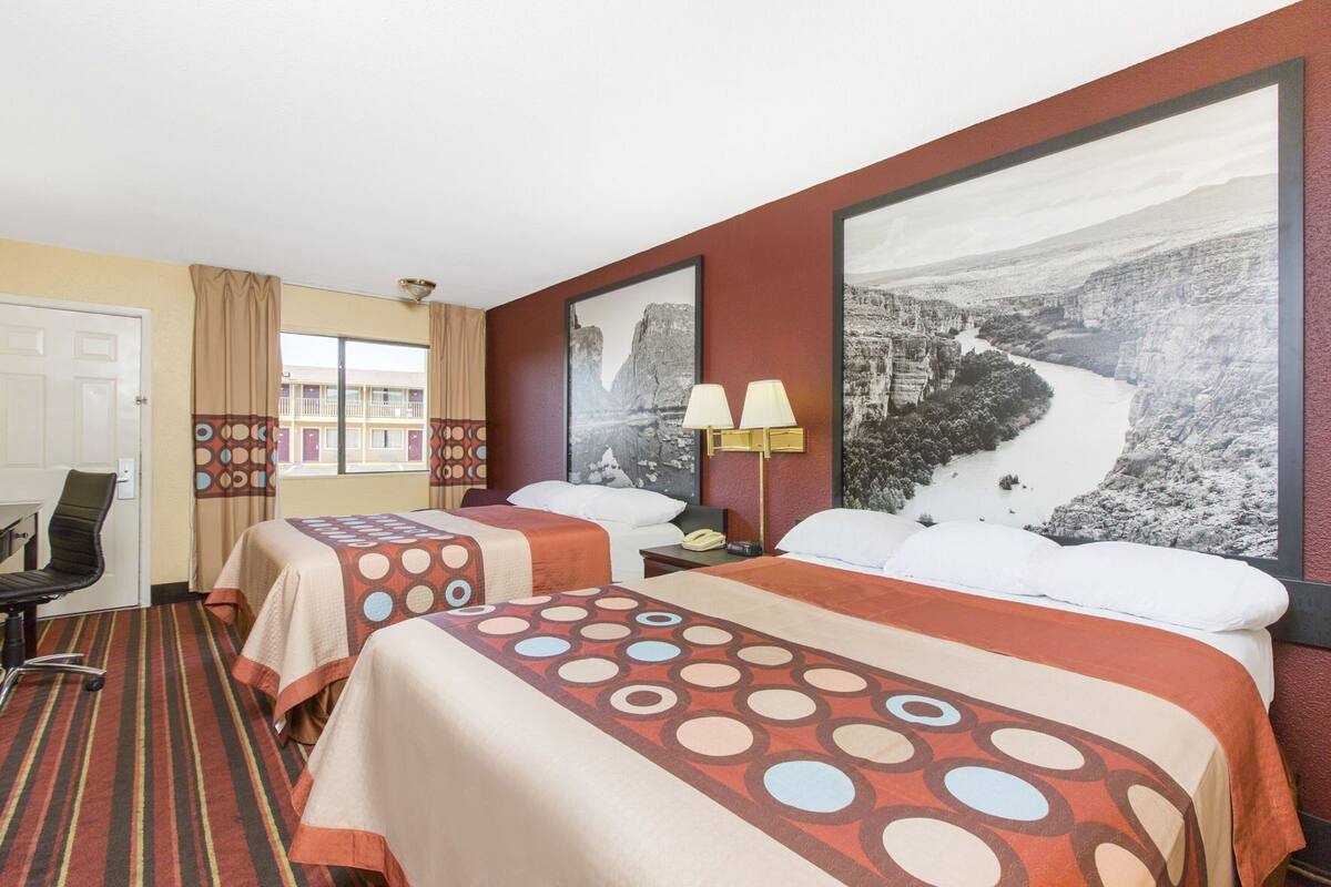 A hotel room with two beds, a desk, a chair, a lamp, and a large window.