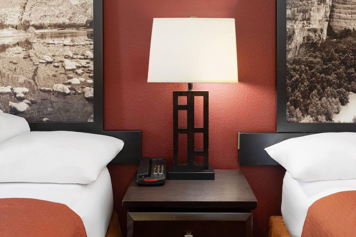 A hotel room with two beds, a nightstand with a lamp, a phone, and framed pictures on the walls.