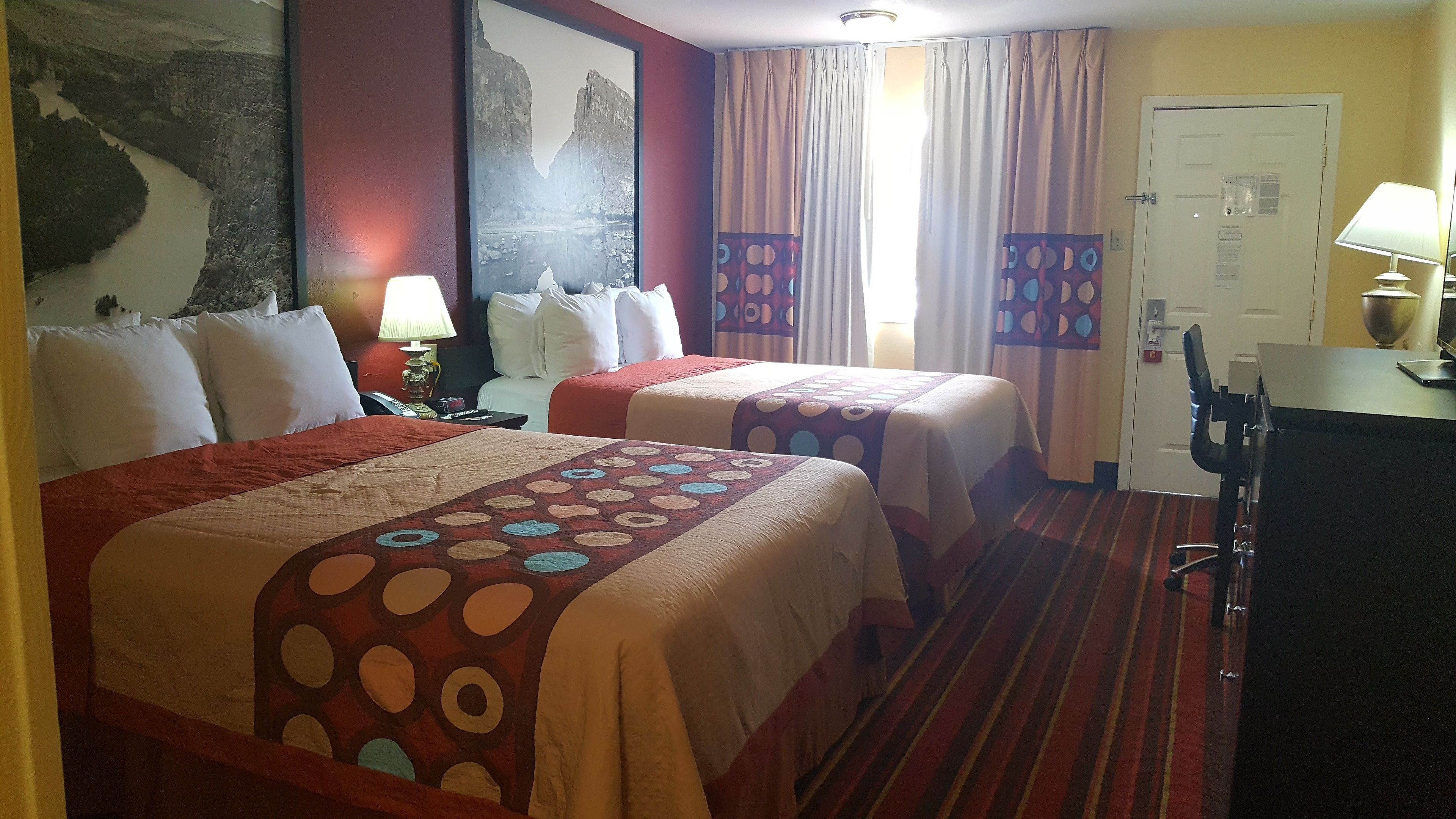A hotel room with two beds, a desk, a chair, a lamp, and a painting on the wall.