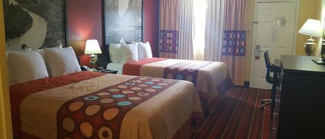 A hotel room with two beds, a desk, a chair, a lamp, and a painting on the wall.