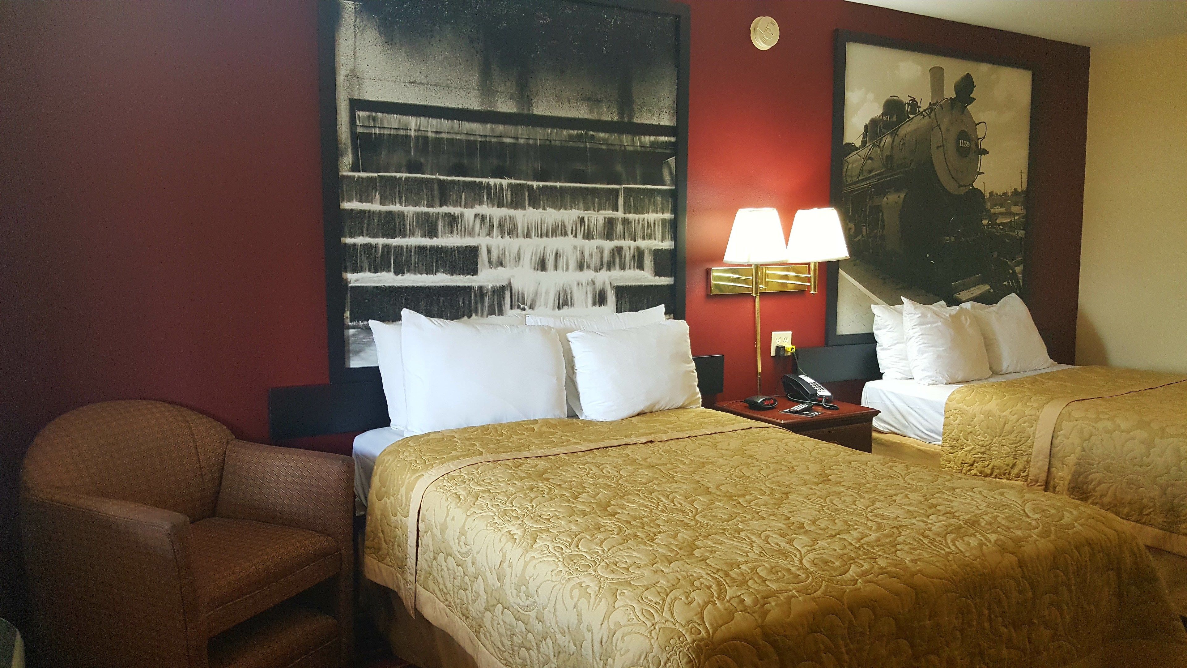 A hotel room with two beds, a desk with a lamp, a chair, and a large abstract painting on the wall.