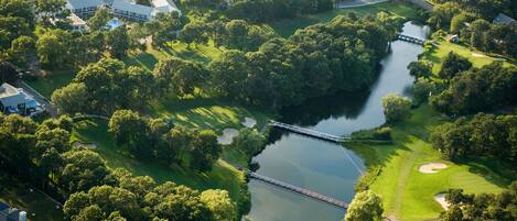 An aerial view of a golf course with a bridge over a lake, surrounded by dense trees and residential buildings.