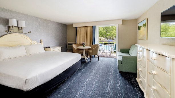 A hotel room with a large bed, a small table with chairs, a green armchair, a dresser, and a balcony with a view of trees.