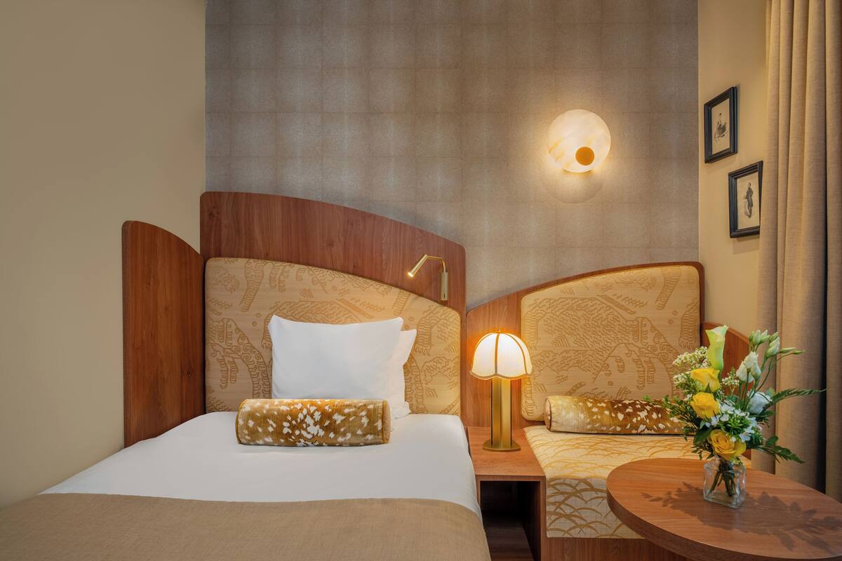 Hotellihuone, jossa on kaksi sänkyä, yöpöytä lampun kanssa, pieni pöytä maljakossa olevilla kukilla ja seinälle asennettu valaisin.